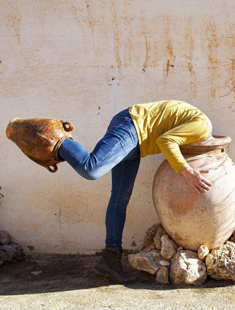 Una persona vestida con una camiseta amarilla y jeans azules está inclinada hacia una jarra de cerámica, pareciendo interactuar con ella mientras su cabeza está fuera de vista.